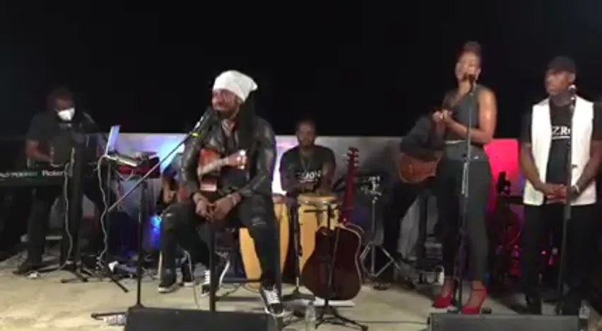 An acoustic ensemble on a night-time stage: a guitarist in a white head wrap, hand drums, keys and backing vocalists