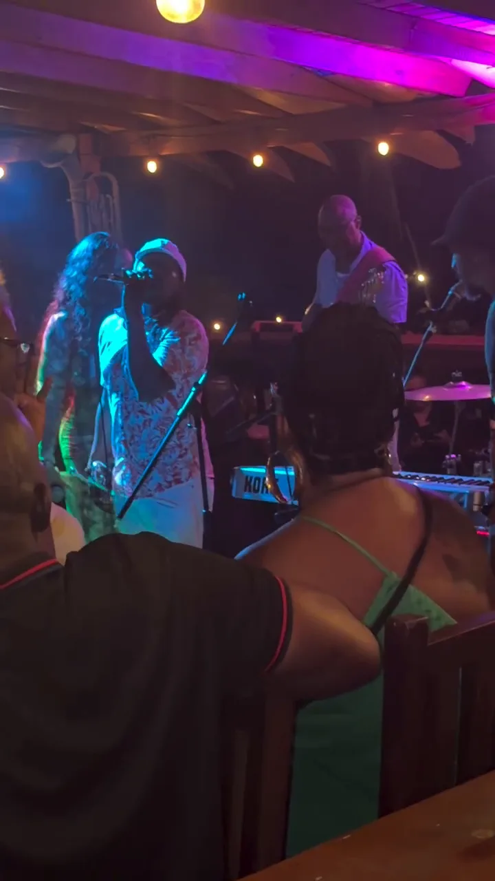 A singer in a white cap performing under purple stage lights with a band, filmed from the audience