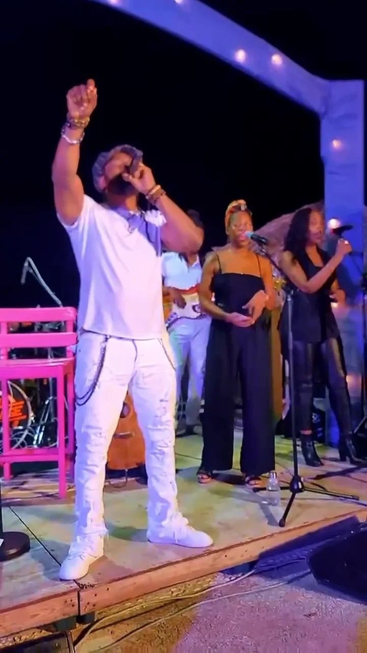 Hezron in white with a raised fist on a wooden stage beneath a blue arch of lights, backing vocalists behind him