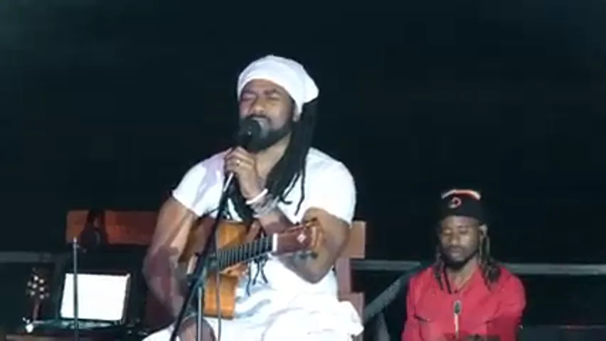 Hezron in white with an acoustic guitar singing at a microphone, a musician in red beside him