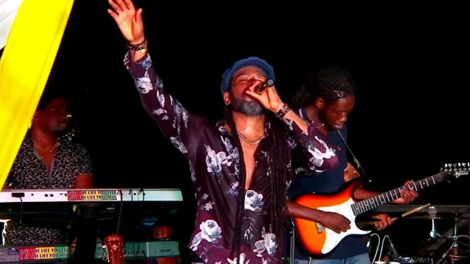 Hezron in a floral shirt and blue cap, one arm raised, singing beside a guitarist with an orange electric guitar