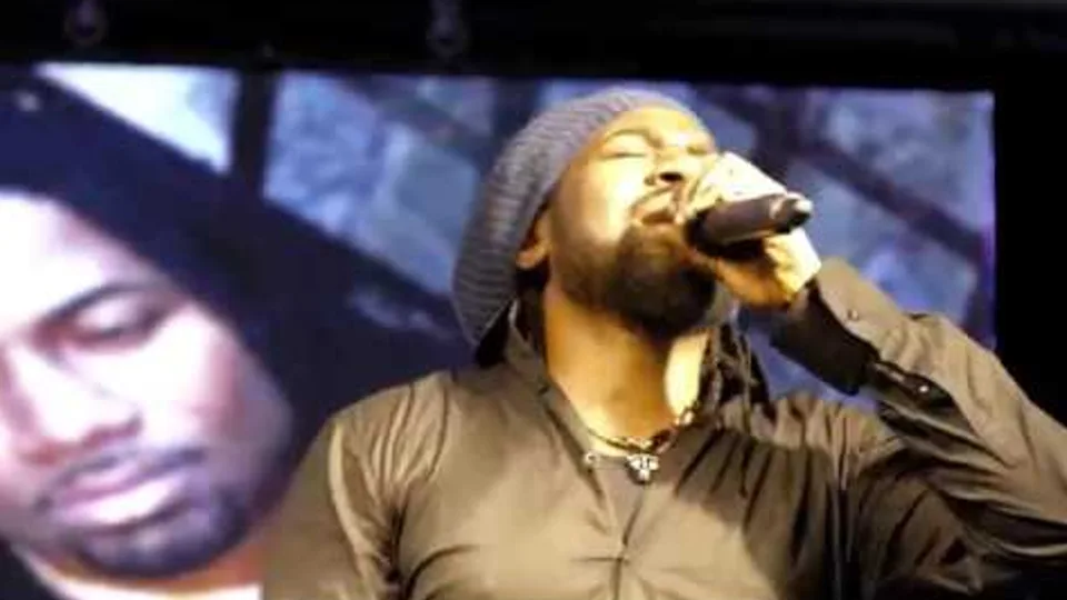 Hezron in a grey beanie singing in front of a giant video screen showing his face